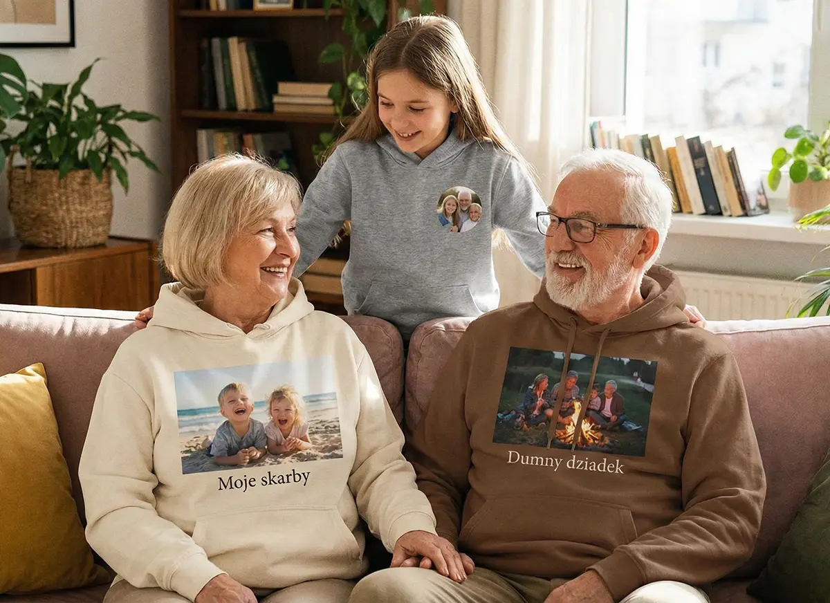Babcia i dziadek siedzą na sofie w bluzach z kapturem ze zdjęciami wnuków i krótkimi napisami, obok stojąca wnuczka.